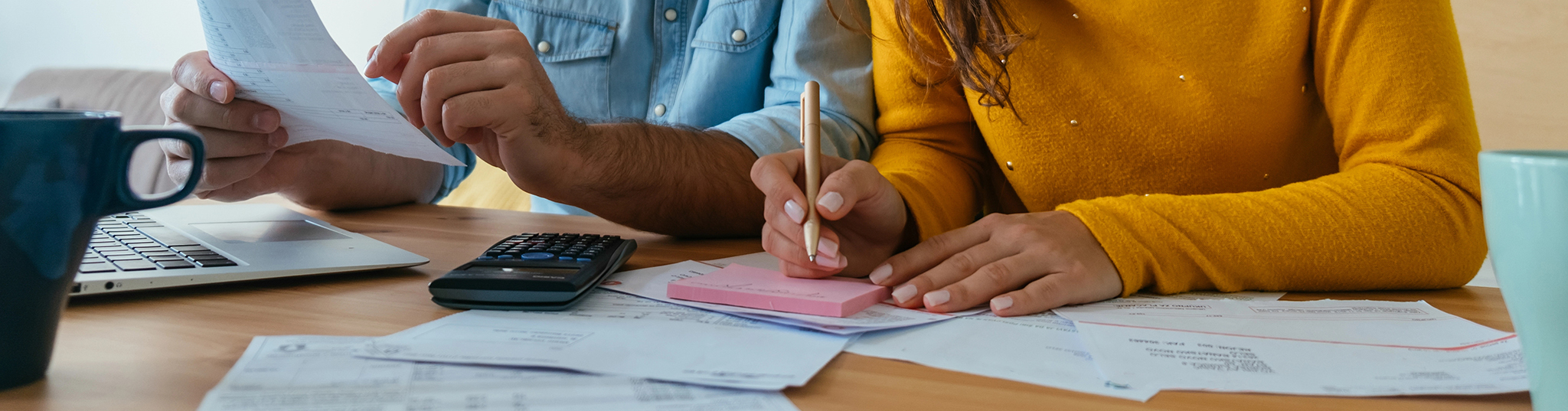 Couple doing finances at desk. Man is wearing blue denim shirt and woman is wearing a gold sweater. Papers, bills and a calculator are on the table. Woman is writing on a sticky note pad with a pen.